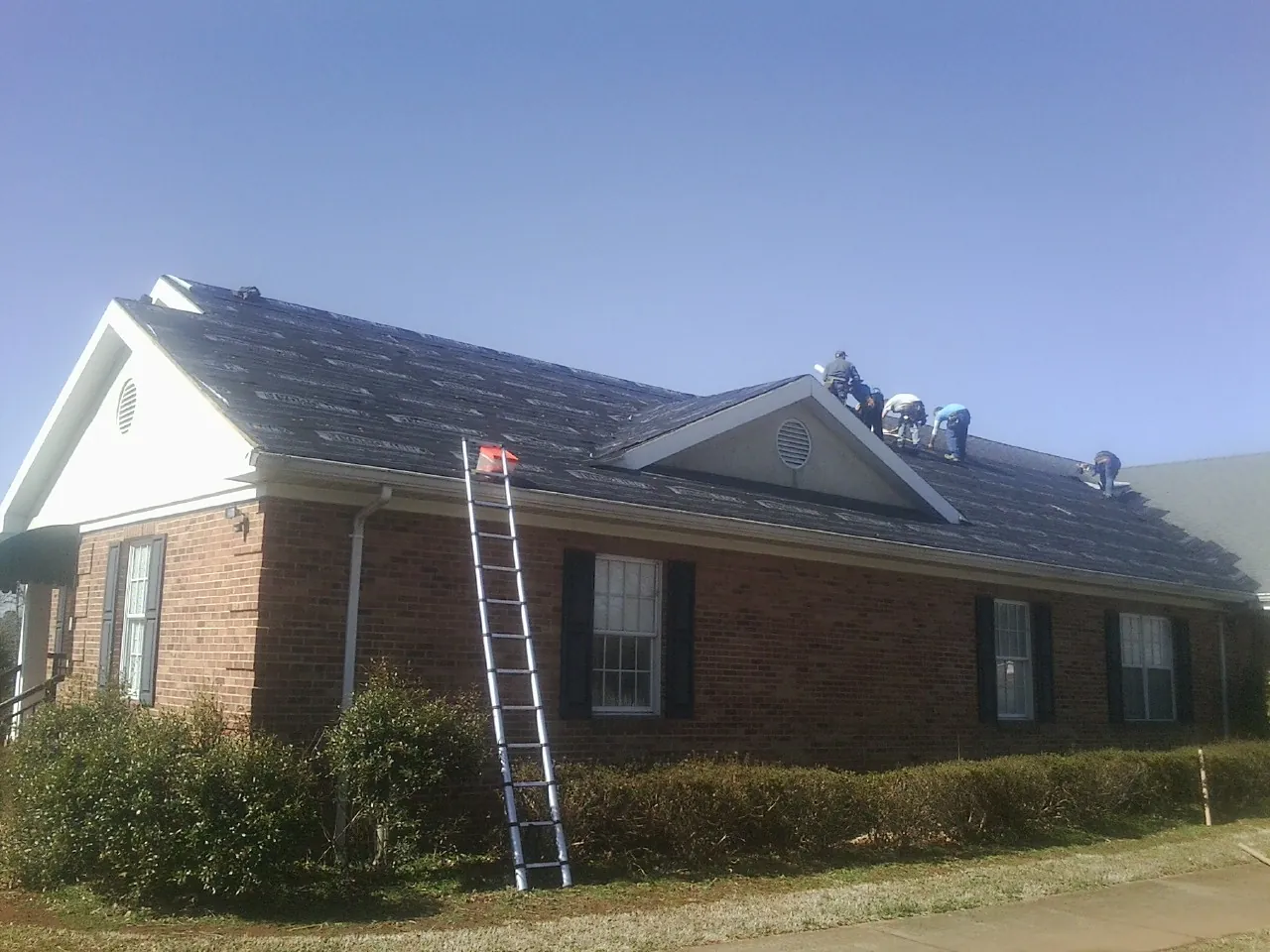 Crew tearing shingles off a brick ranch roof with a ladder set — Emergency Roof Repair work by our crew in Ponsford