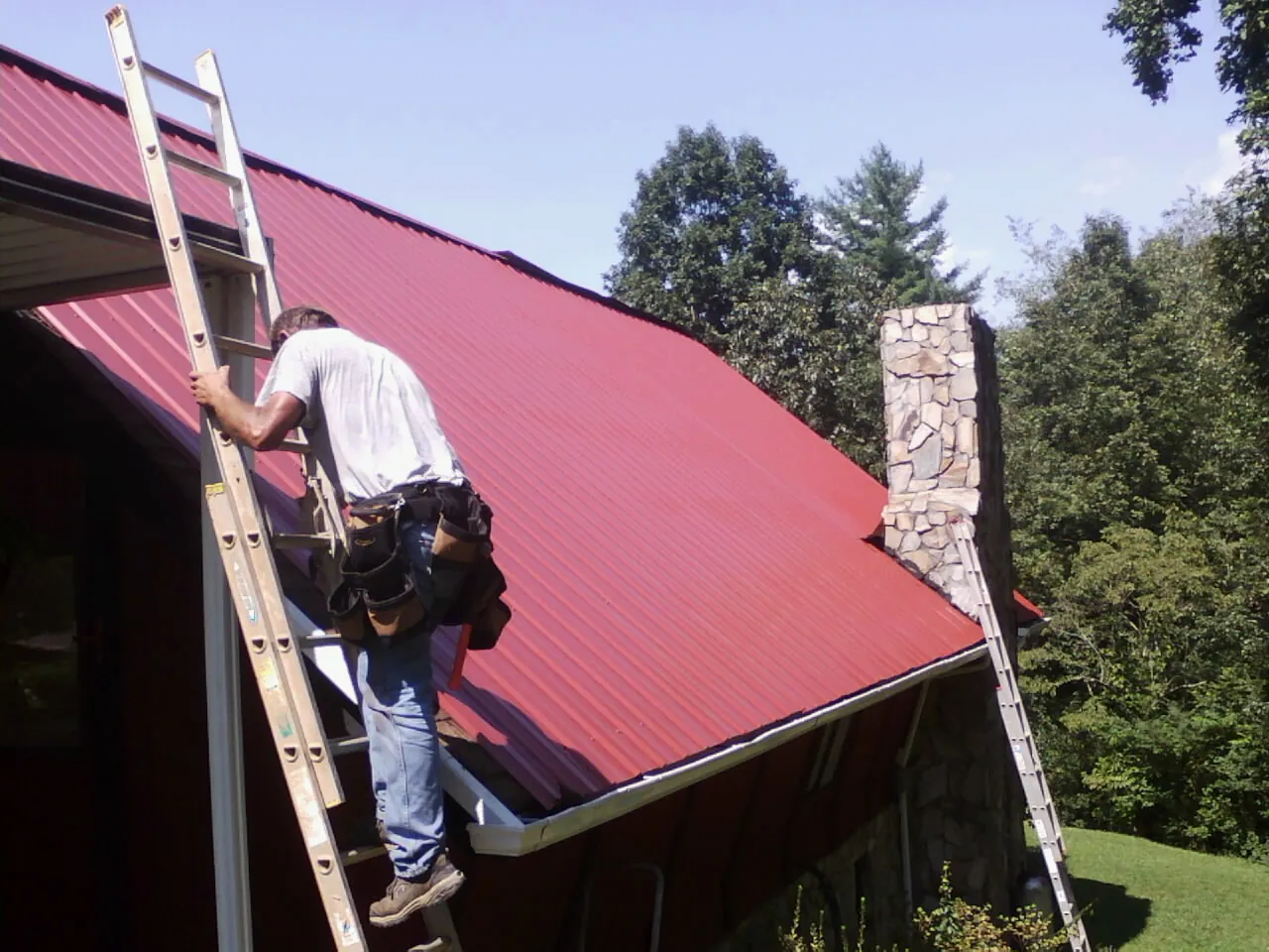 Roofer on a ladder beside a red metal roof and stone chimney — Soffit & Fascia Repair by Roofing Brothers in Ponsford